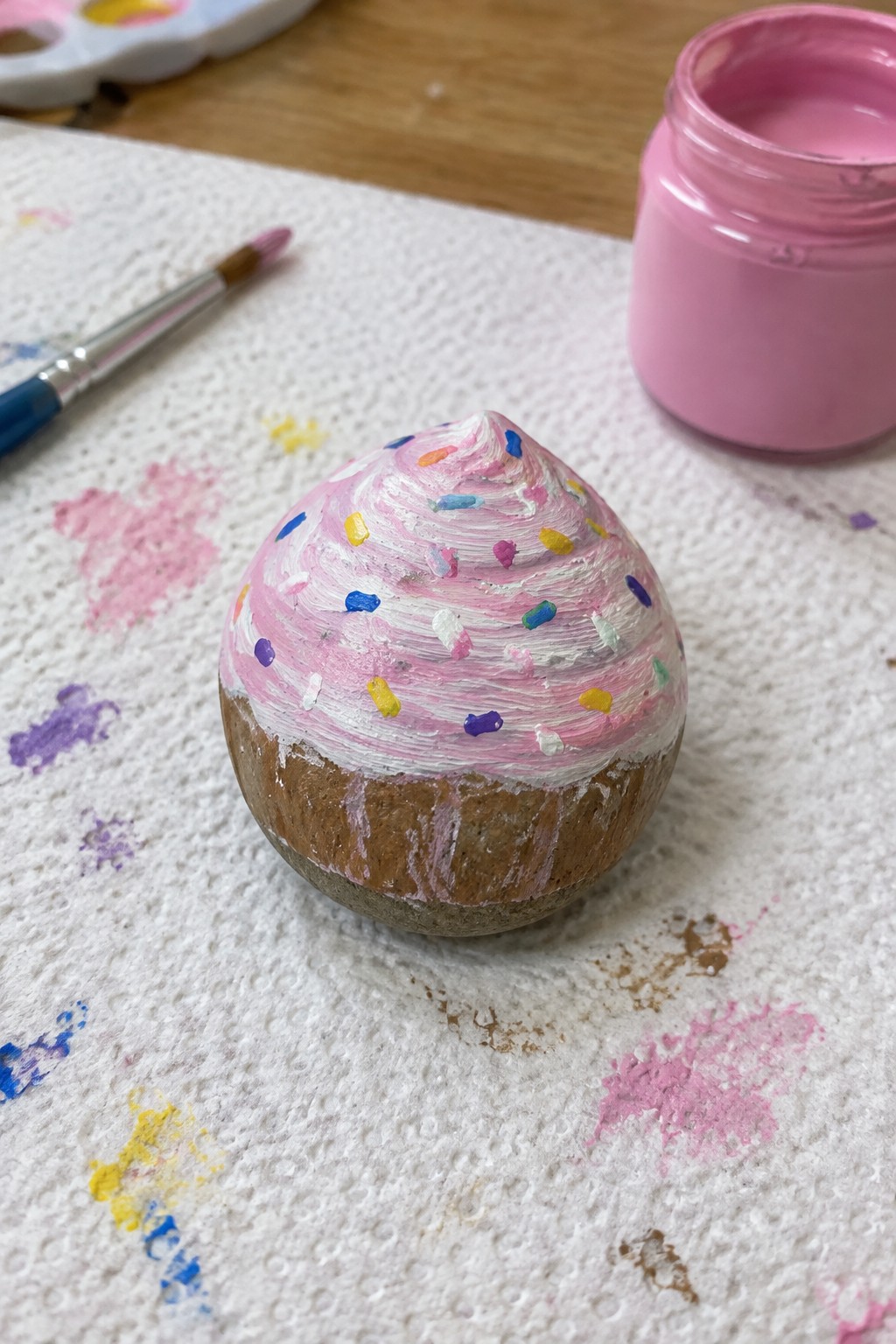 Painted rock cupcake with pink swirled frosting and colorful sprinkles on paper towel