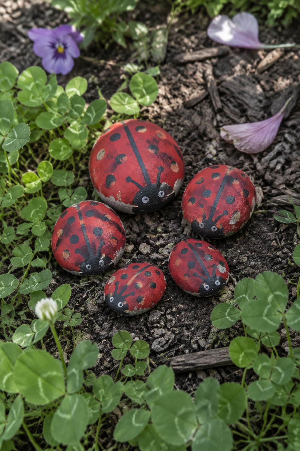 Five red painted ladybug rocks amid green plants, soil, and purple flowers