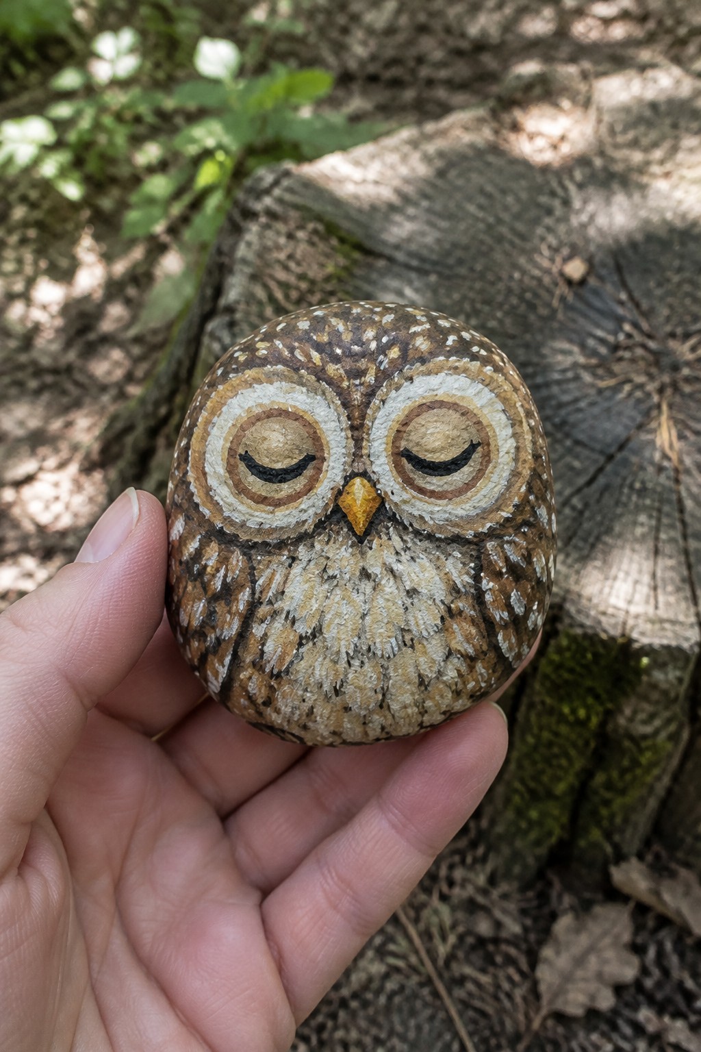 A hand holds a small round rock painted as a sleeping owl with brown feathers, white accents, closed eyes, and a yellow beak.