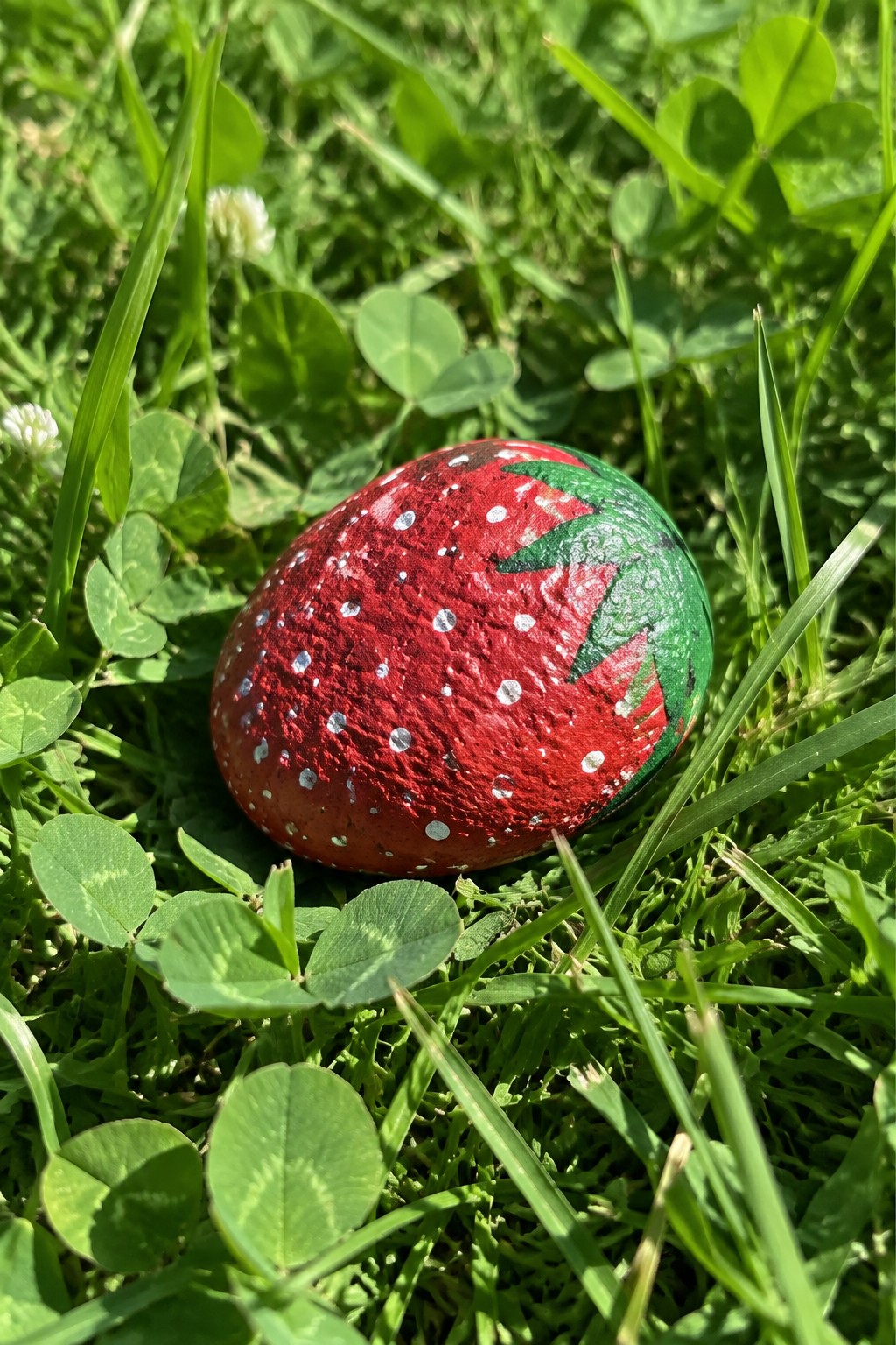 Painted strawberry rock with red body, white dots, and green leaves on grass