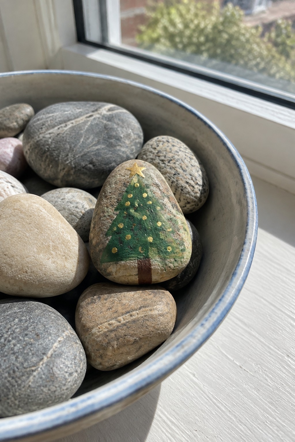Bowl of smooth stones on a windowsill, one painted as a Christmas tree.