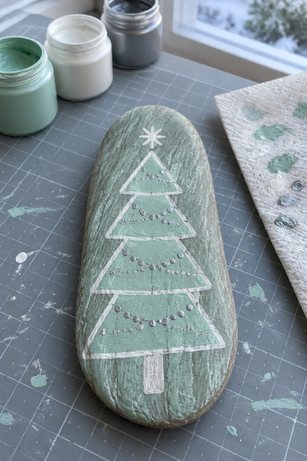 Painted green Christmas tree rock with white outline, star and silver dots on mat