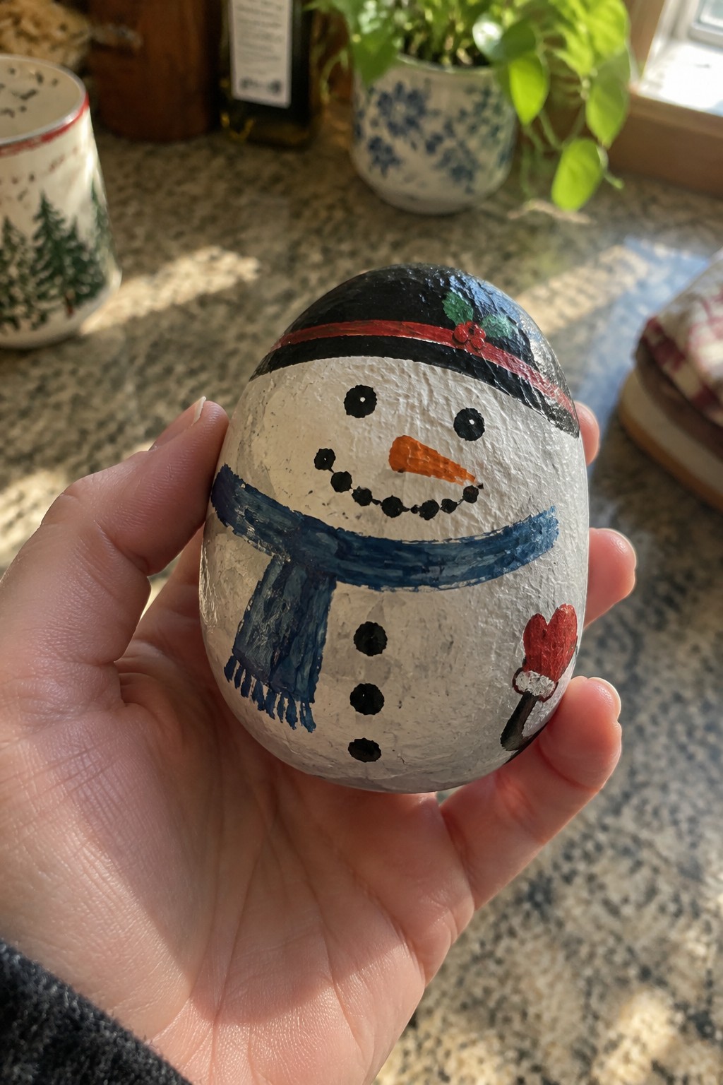Hand holding painted snowman rock with black hat, blue scarf, and orange nose.