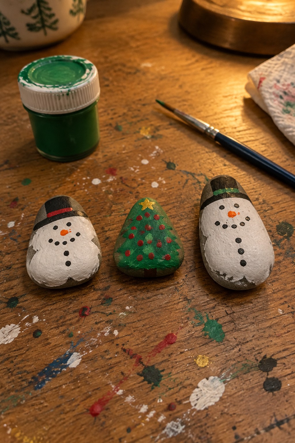 Two snowmen and Christmas tree painted on rocks amid supplies on splattered wood