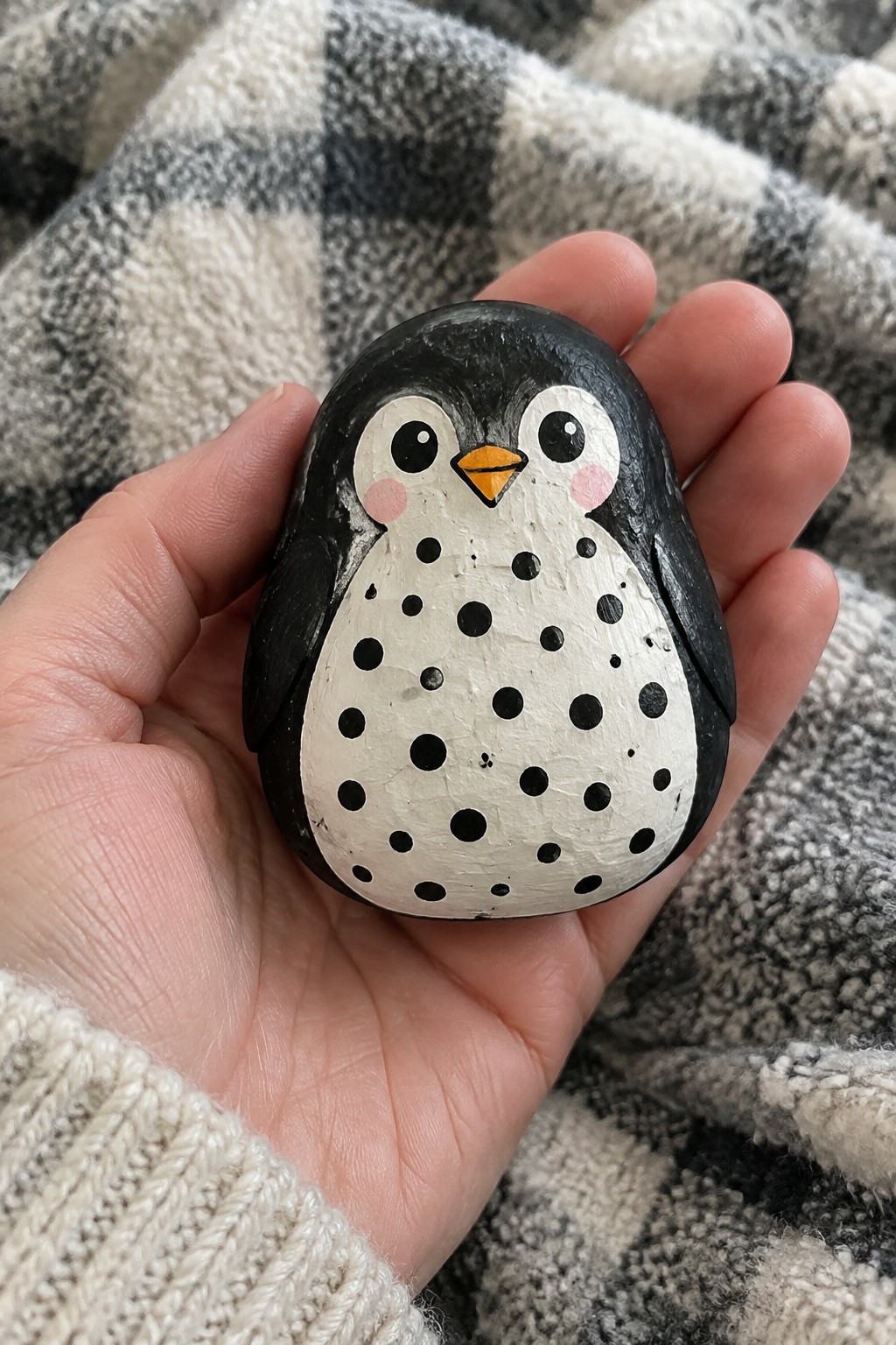 Hand in cream sweater holding black-and-white spotted penguin rock on plaid blanket