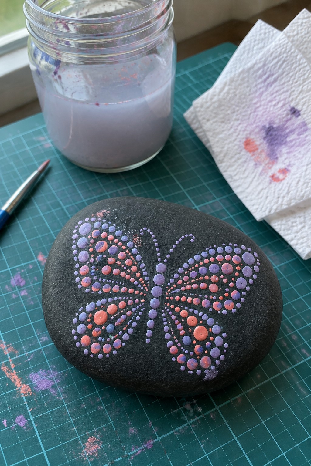Black rock with dotted butterfly in purple and coral on green mat beside paint jar