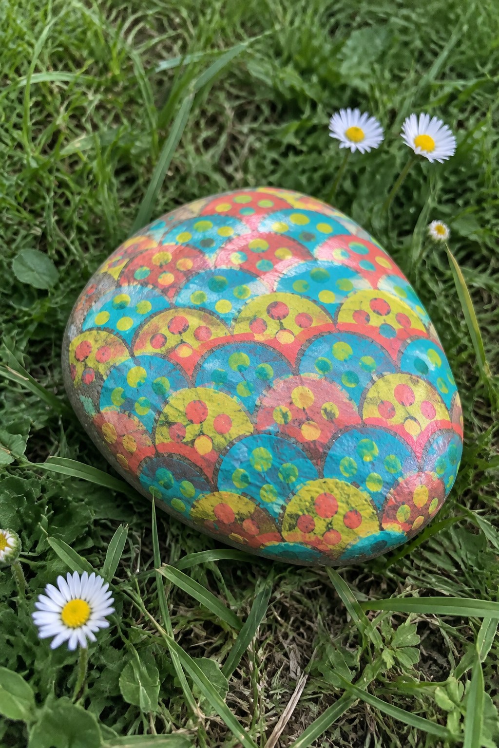 A rounded stone painted with colorful overlapping semicircles filled with dots sits in grass among daisies.
