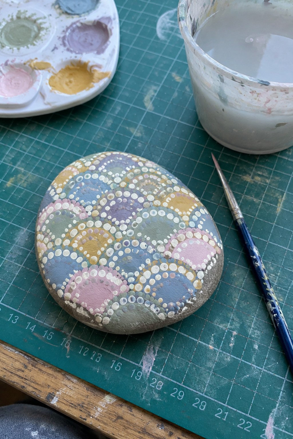 Painted stone with pastel dotted circles beside palette, brush, and water cup on green mat