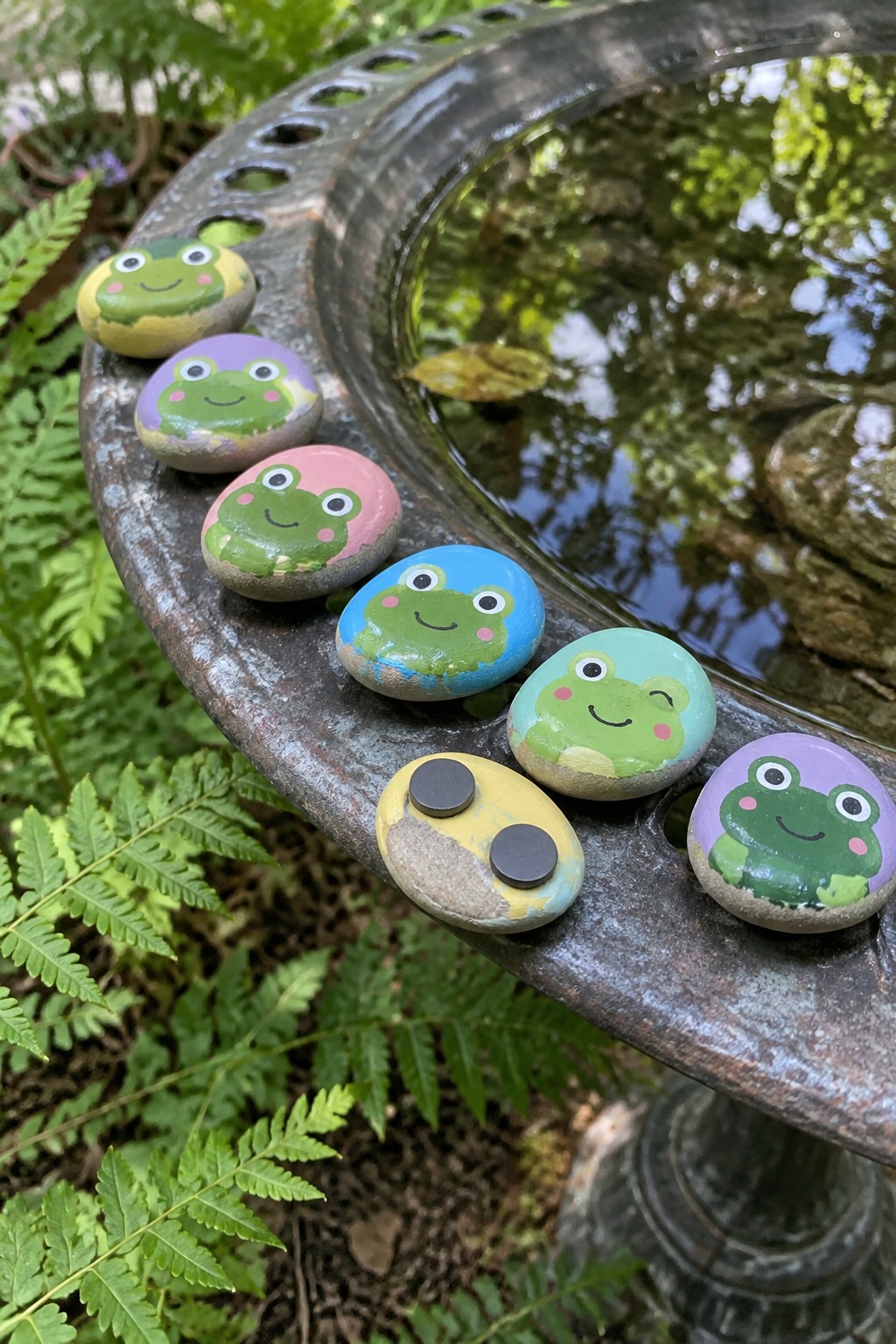 Colorful hand-painted frog rocks lined along a weathered garden fountain rim