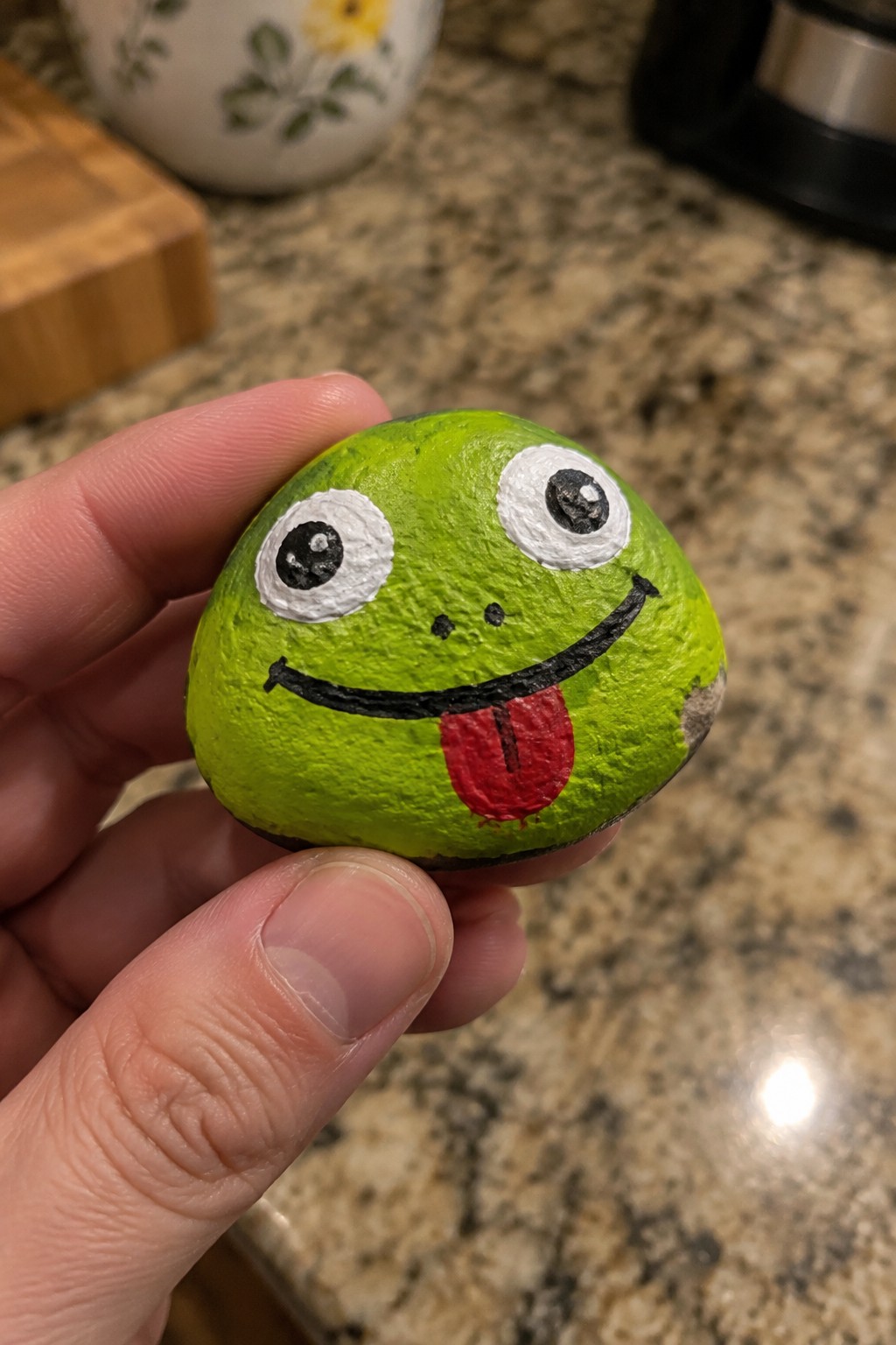 Hand holding lime-green rock painted as cartoon frog with big eyes and red tongue