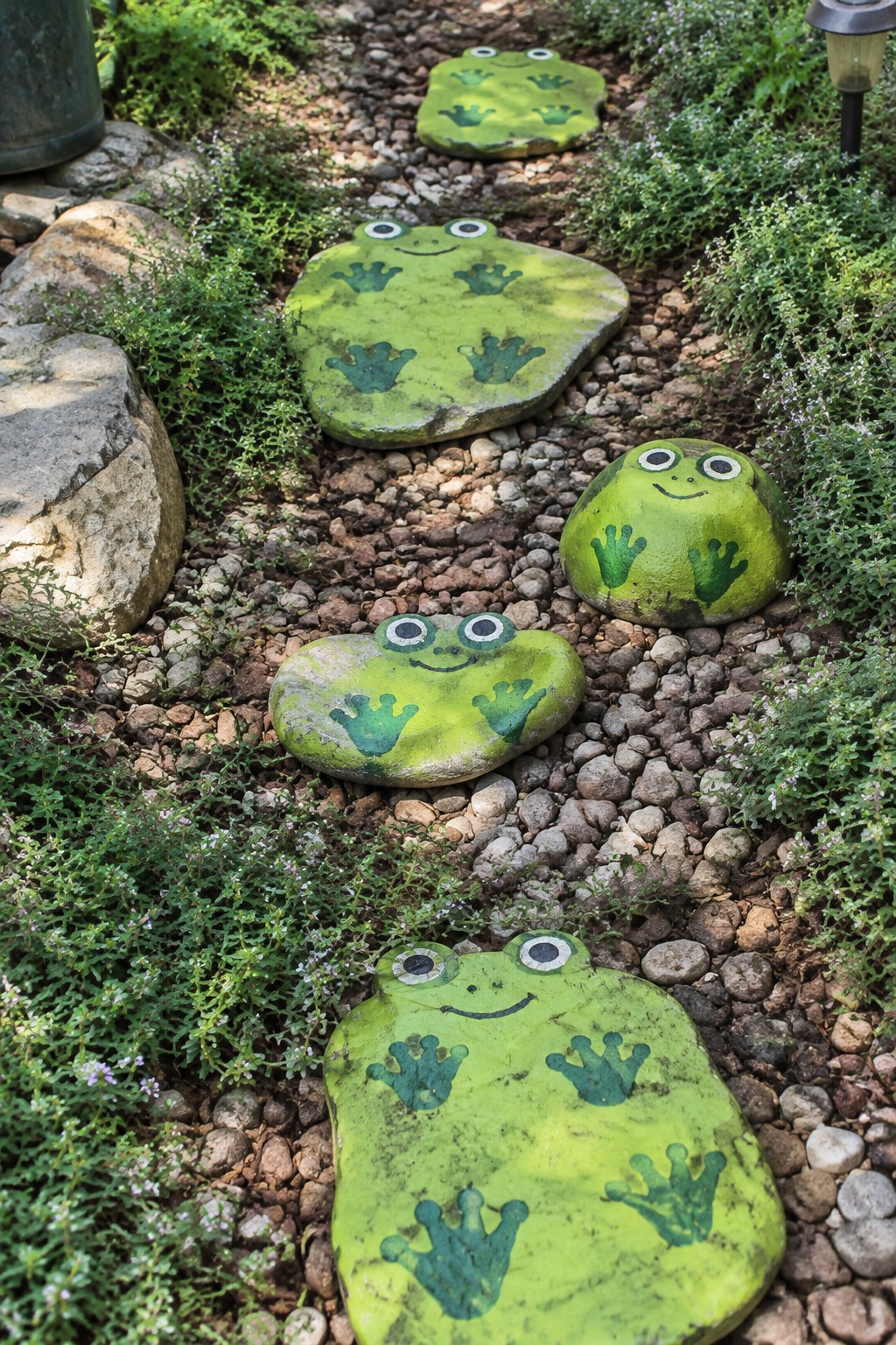 Green frog-shaped rocks with handprints and smiling faces on garden gravel path