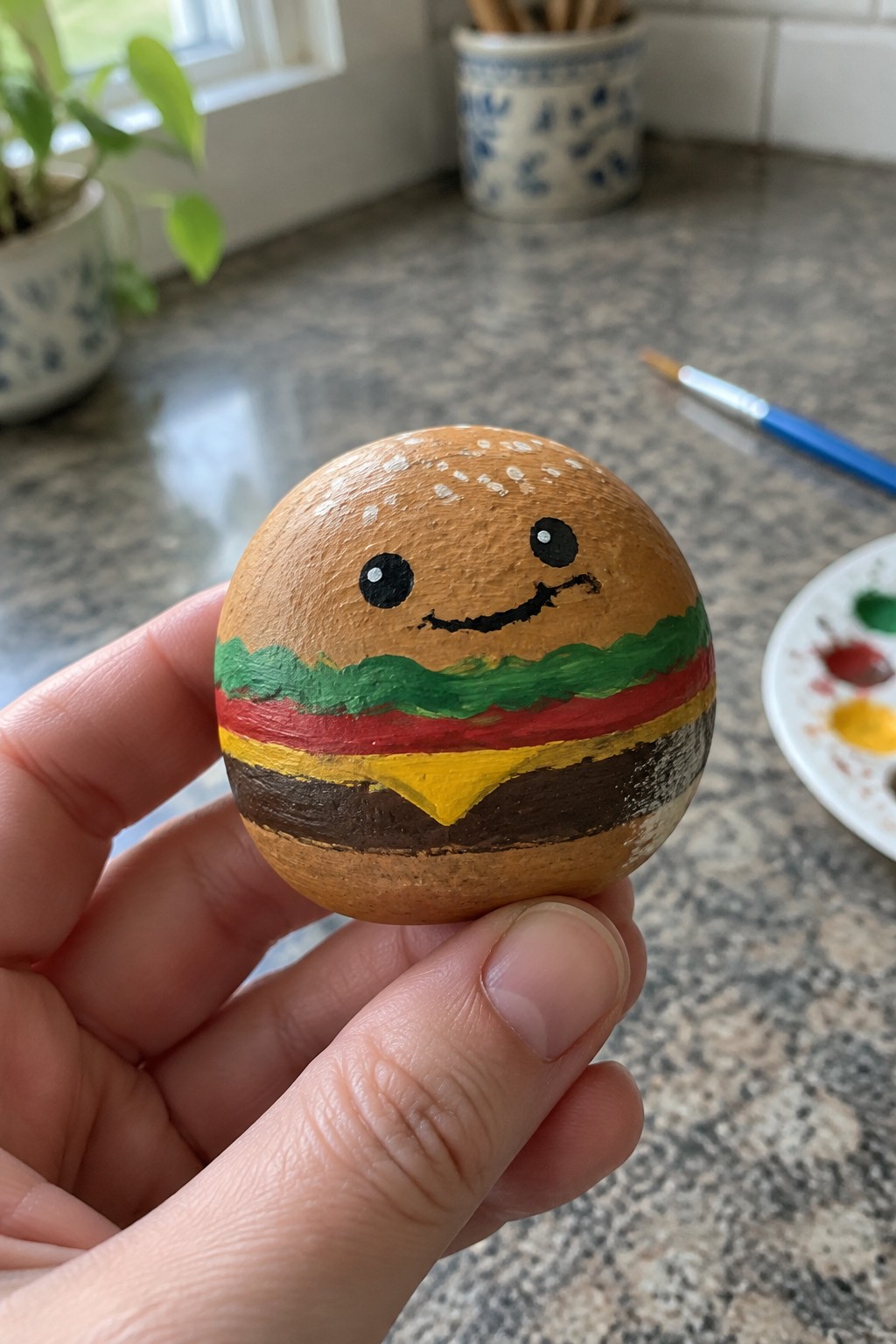 Hand holding smiling hamburger rock painted with sesame bun, lettuce, and cheese layers.