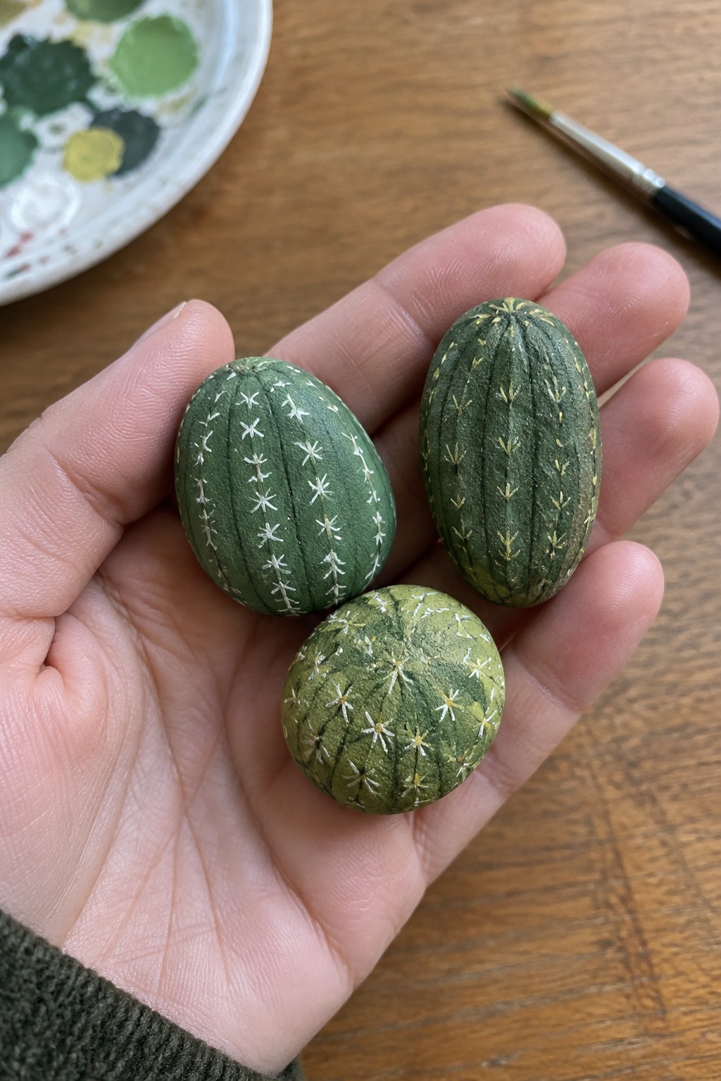 Hand holding three green painted cactus rocks near palette and brush on wood
