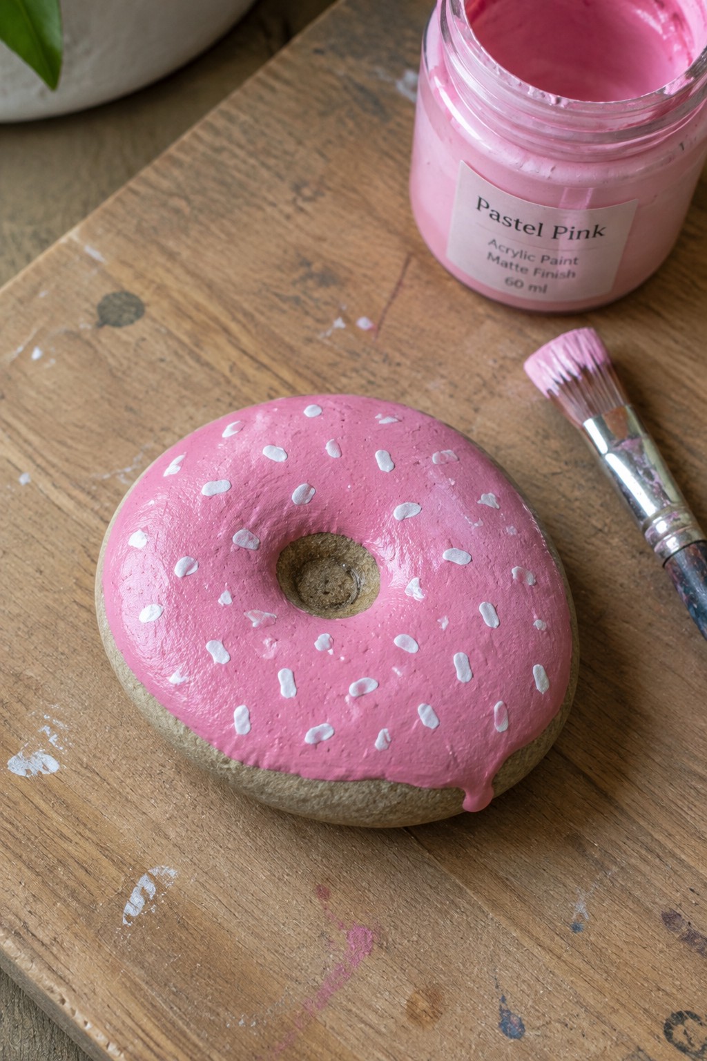 Pink painted donut with white sprinkles beside pastel pink paint jar and brush on wood.