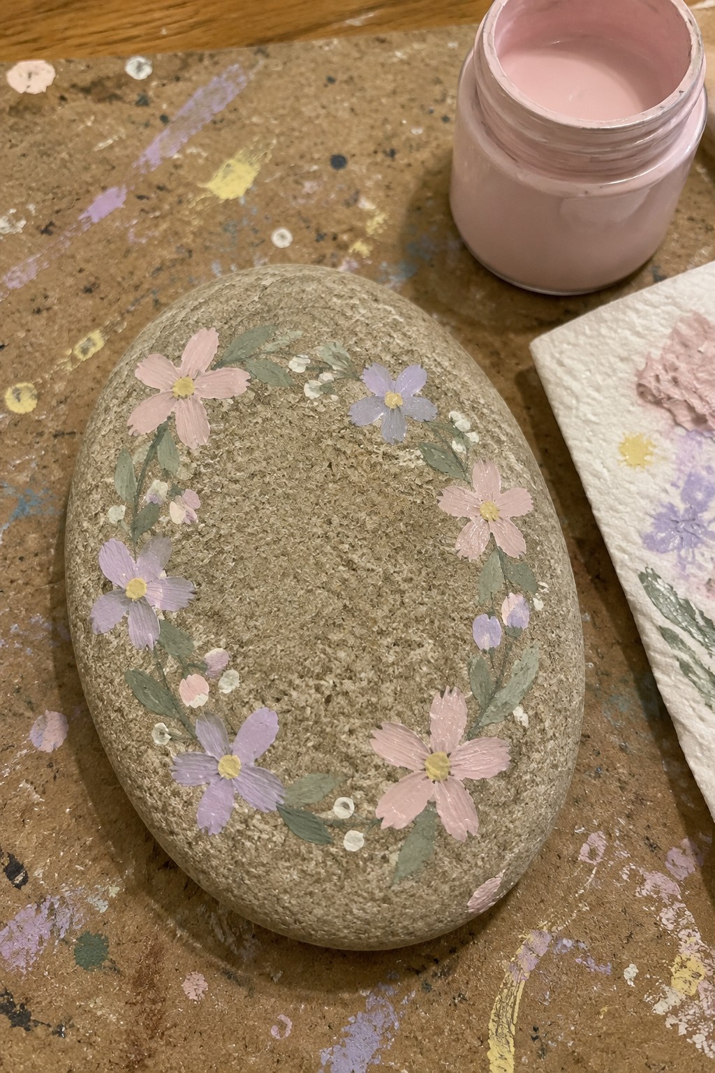 Painted stone with pink-purple floral wreath beside open paint jar on splattered table