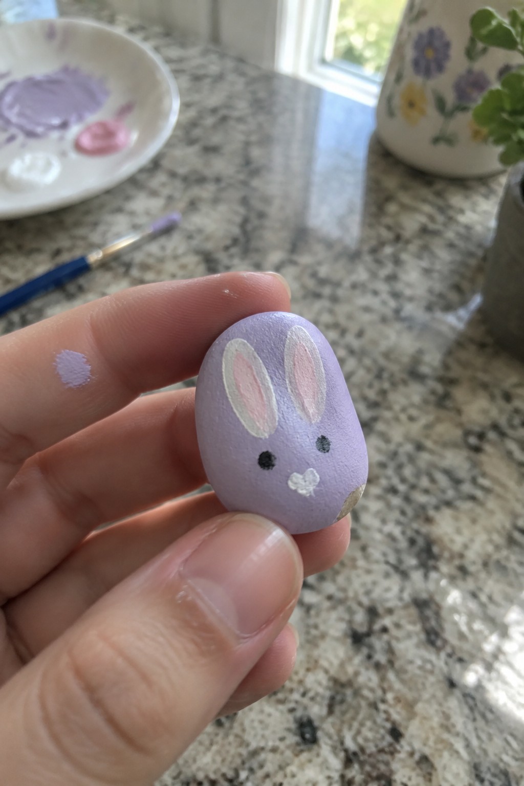 Hand holding purple painted stone bunny with pink inner ears on marble counter