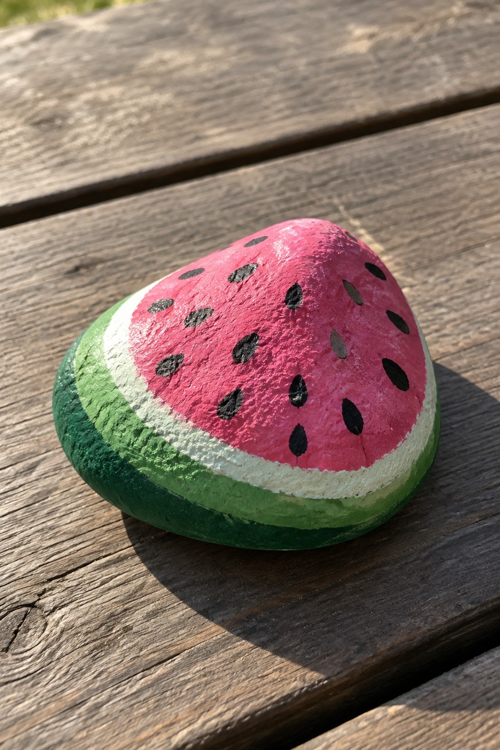 Painted rock resembling watermelon slice with green rind and pink seeds on wood.