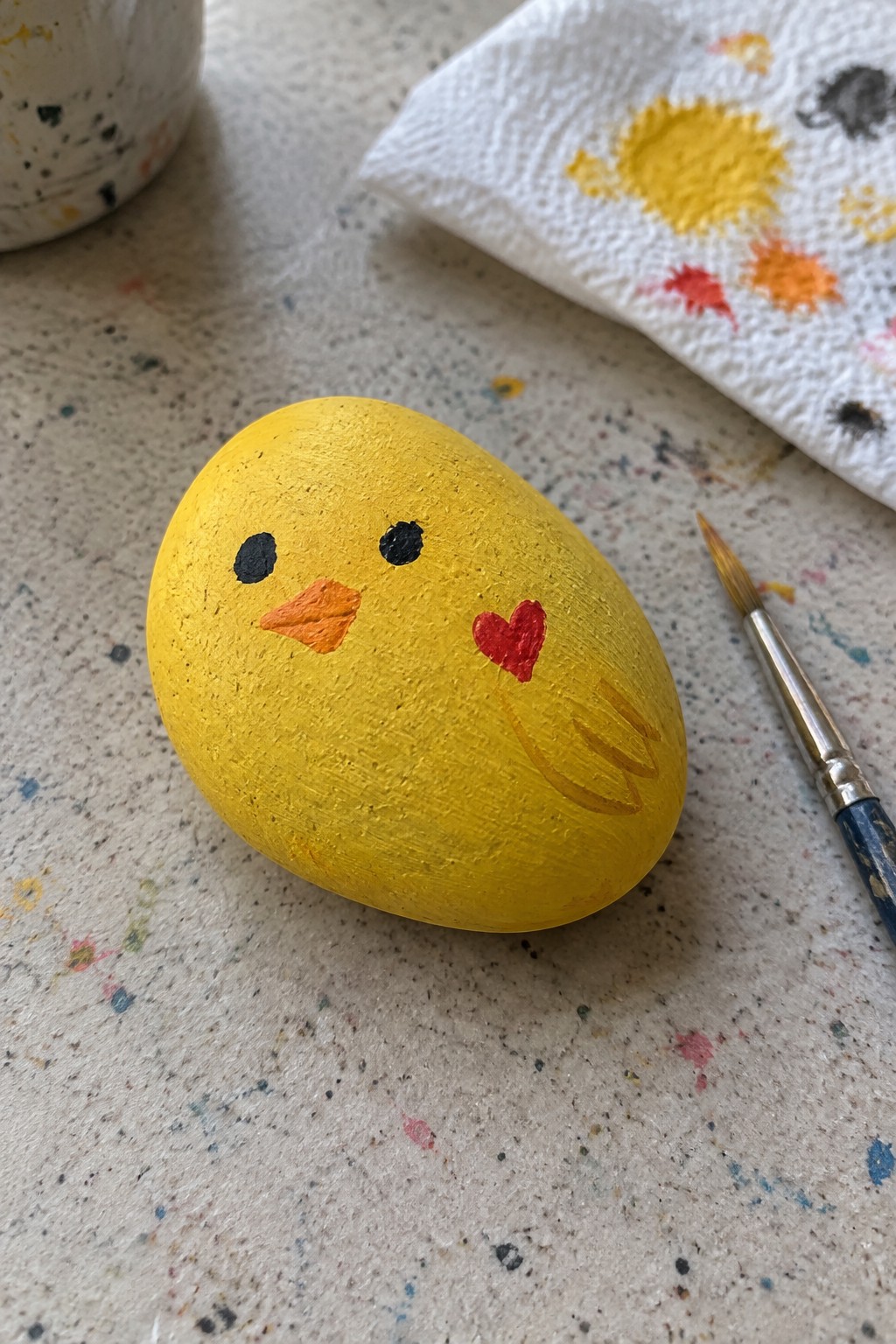 Yellow chick rock with black eyes, orange beak, red heart beside paintbrush