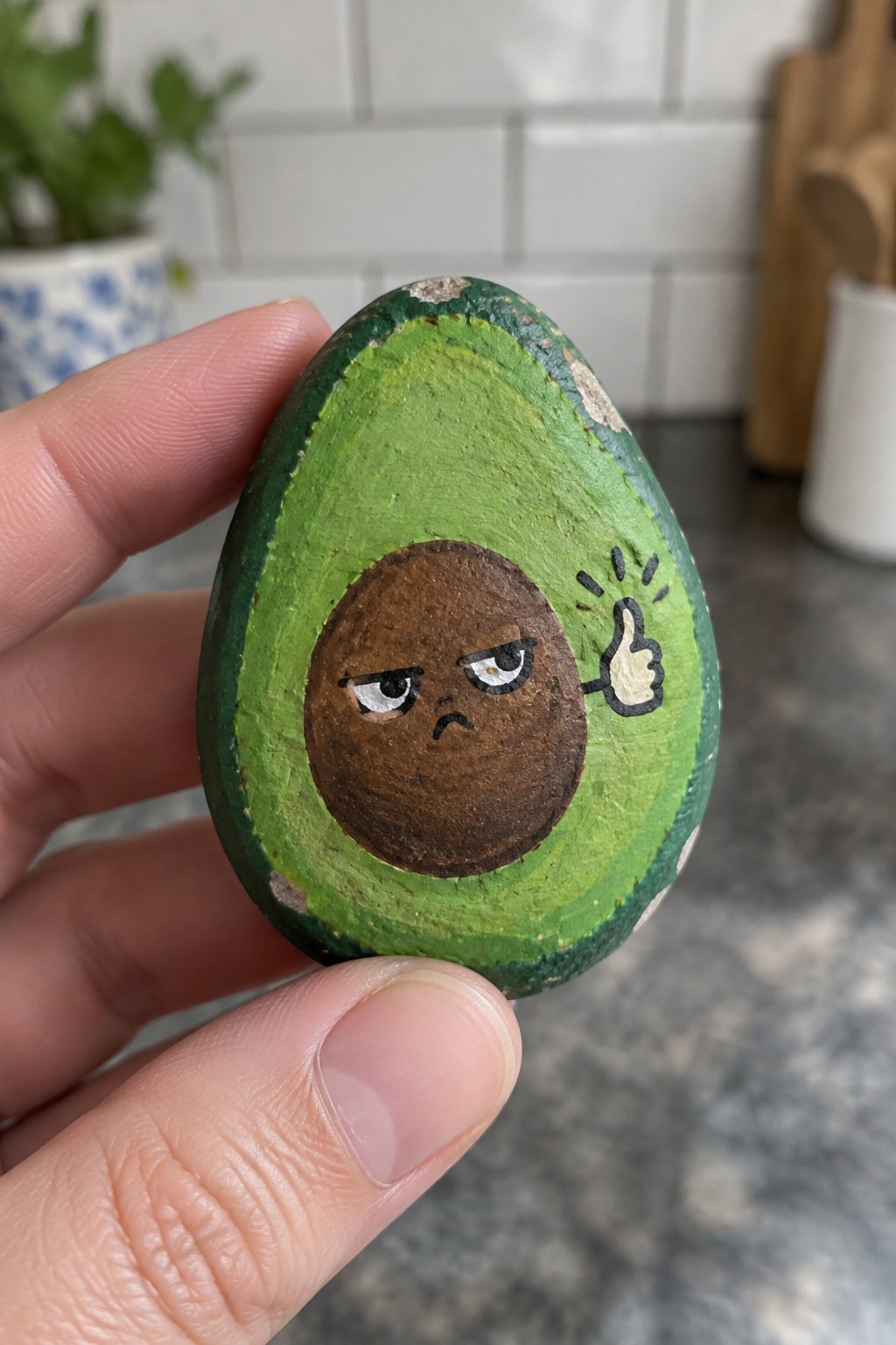 Hand holding painted avocado rock with grumpy cartoon pit giving thumbs-up gesture