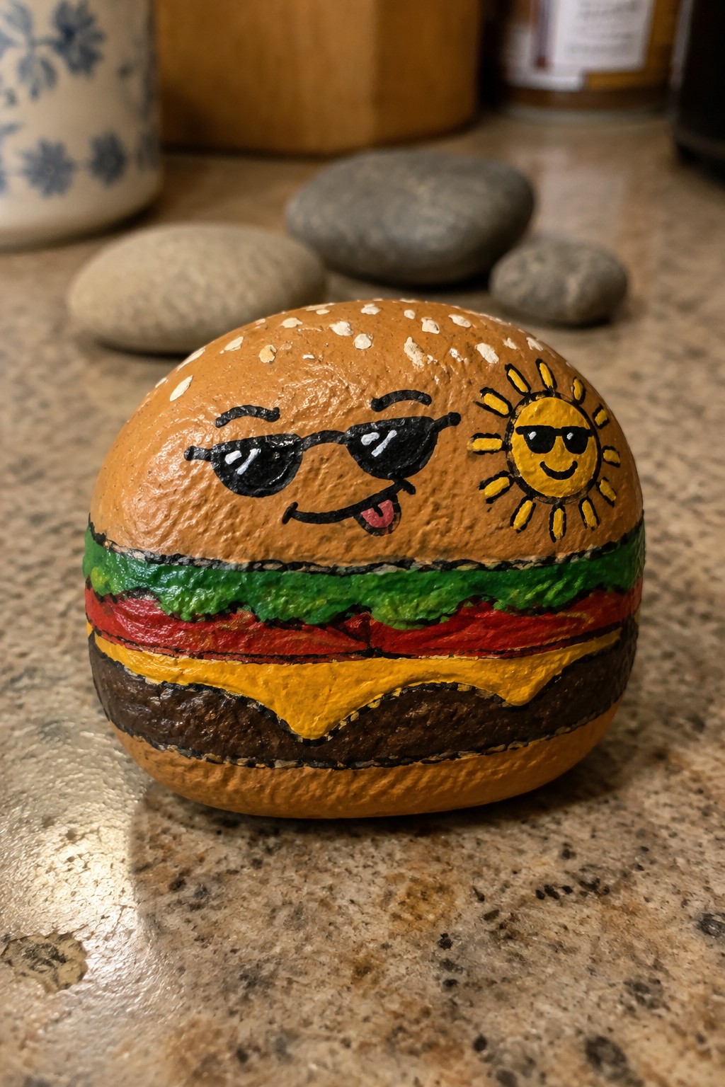 Painted rock hamburger with sesame bun, cartoon sunglasses face, smiling sun, and layered fillings.