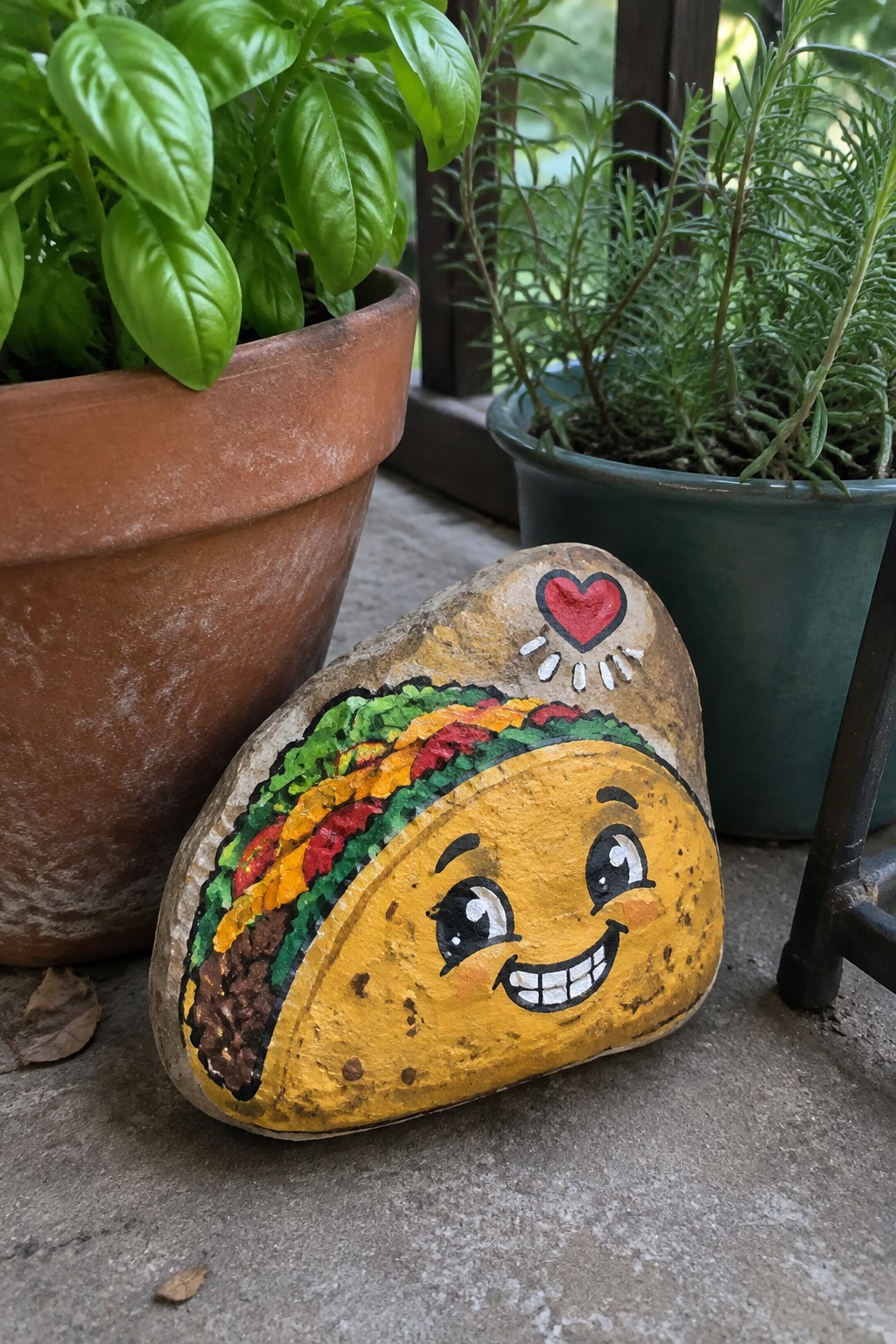 Painted smiling taco rock with heart between basil and rosemary potted plants