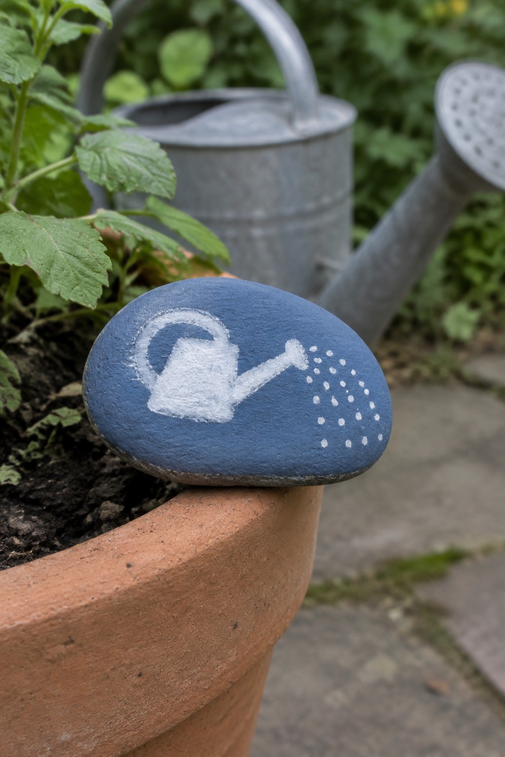 Blue rock with white watering can painted on terracotta pot rim.